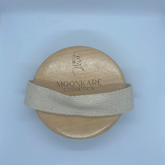 Brosse exfoliante en bois avec le logo de Moonkare Cosmetics, maintenue par une lanière en tissu naturel.